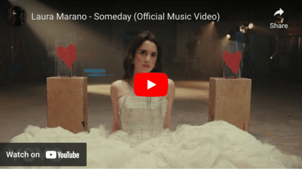 A screenshot from Laura Marano's "Someday" music video, featuring her sitting in a white dress with a despondent expression, surrounded by cardboard cut-outs with hearts, ready for video