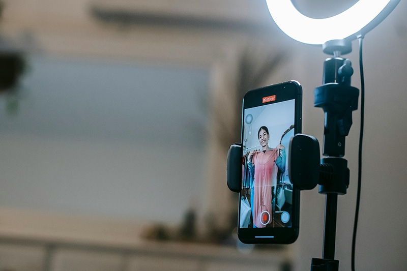 A person holding a pink dress stands in front of a smartphone mounted on a tripod with a ring light, capturing the perfect shot to share their latest fashion tips, much like sharing how to Get Your Music On YouTube—showcasing creativity in every frame.