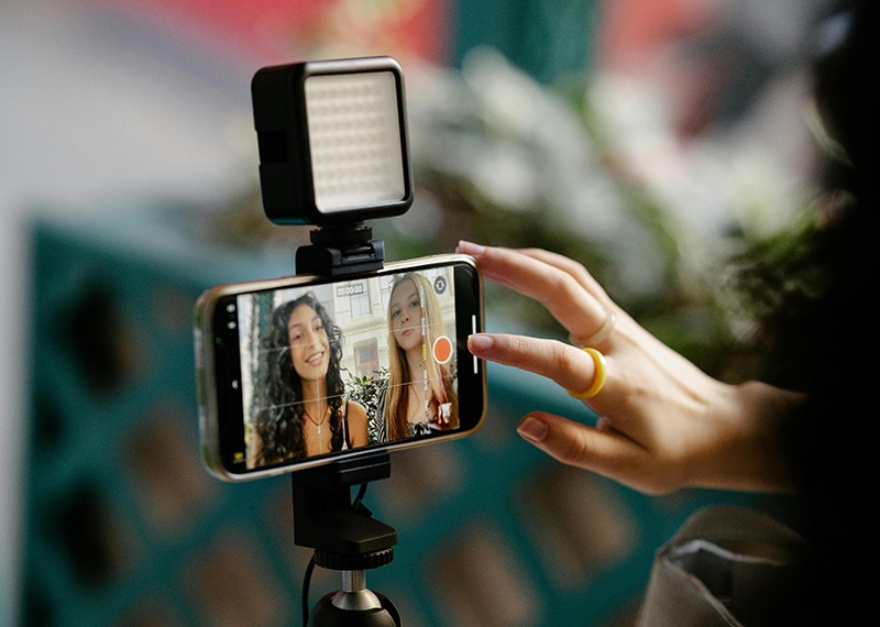 Two people are recording a video on a smartphone mounted on a tripod with a light attached, preparing to get their music on YouTube. One person adjusts the phone while the screen shows their live image, ensuring everything is perfect for their online debut.