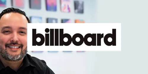 A man smiles in an office with colorful framed art on the wall; the Billboard logo is prominently displayed in the foreground, suggesting a lively press environment.