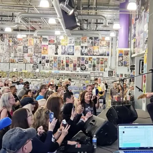 A musician performs on stage with a guitar to a seated audience inside a record store decorated with posters and CDs, celebrating the era of physical music distribution.