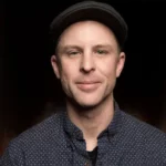 A man wearing a black cap and patterned button-up shirt smiles at the camera against a dark background.
