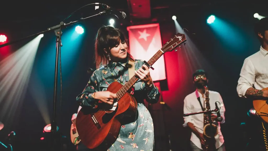 A woman plays an acoustic guitar on stage with a saxophonist and other musicians under colorful lights, while a flag featuring a star hangs in the background—capturing a perfect moment for Music Analytics.