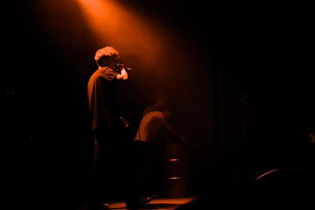 A person with a microphone performs on a dimly lit stage under an orange spotlight, capturing the energy that Music Analytics could quantify, while another figure stands in the background.