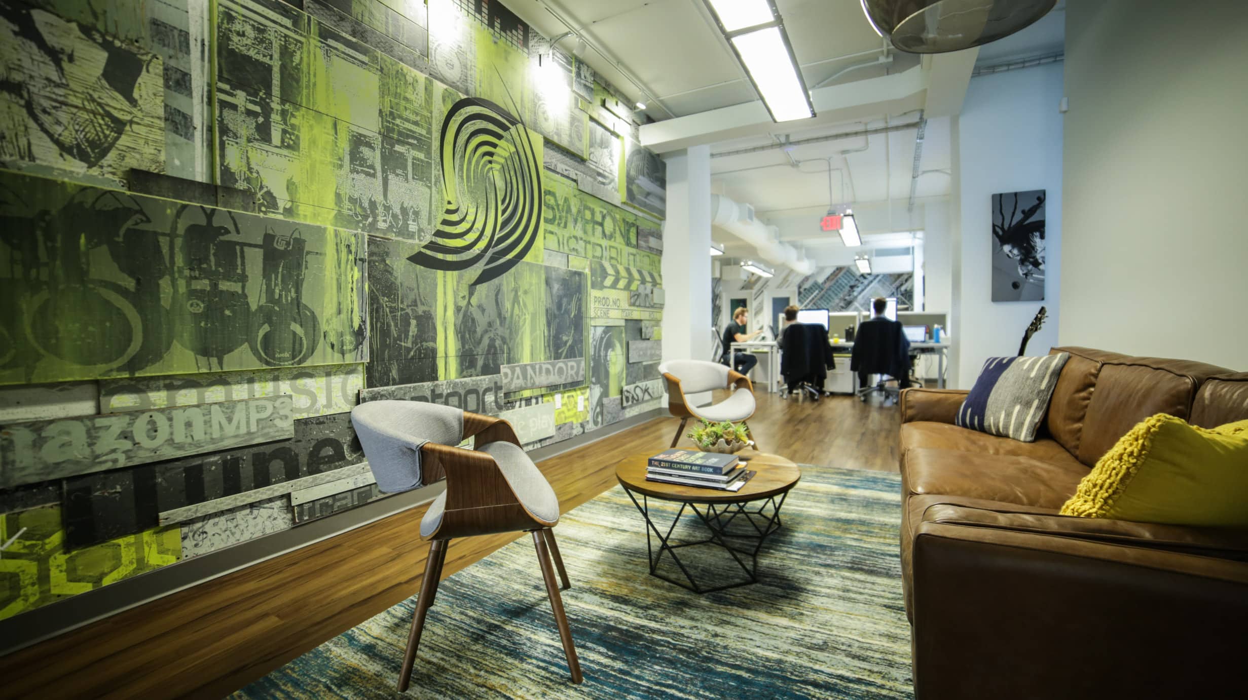 Modern office lounge featuring a brown leather sofa, two gray chairs, a rug, and coffee tables, set against Symphonic NEXT’s signature green and black graphic art; an office area is visible in the background.