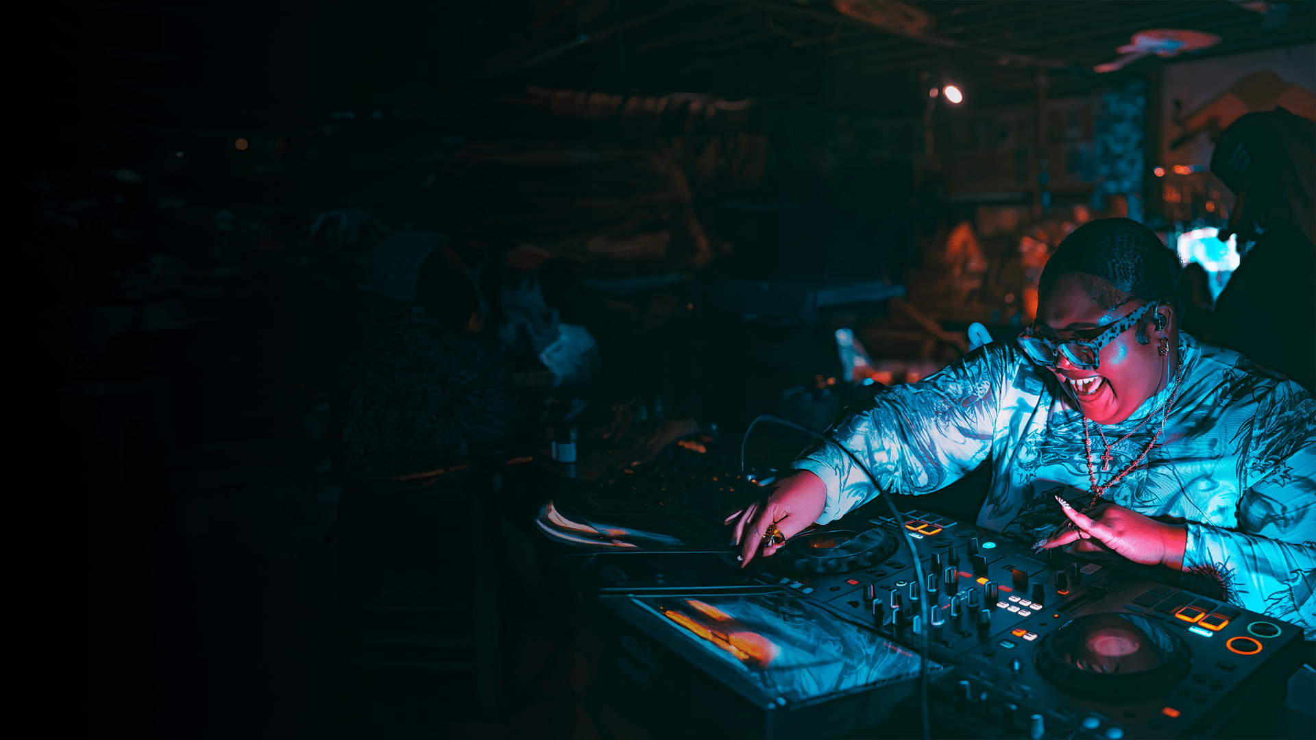 A DJ wearing sunglasses and a patterned shirt is smiling while mixing music on turntables in a dimly lit venue.