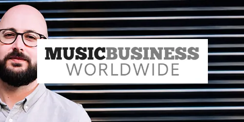 A man with glasses and a beard stands beside a "Music Business Worldwide" press sign in front of a black striped background.