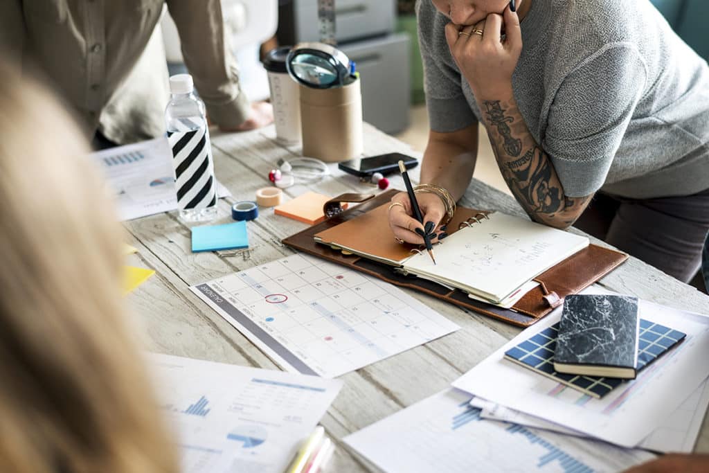 Person writing in a planner at a desk covered with documents, charts on royalty advances, a calendar, notebook, water bottle, and office supplies.