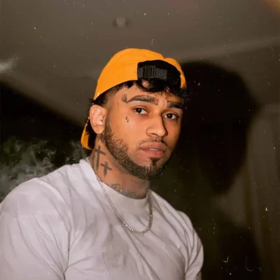 A man with facial tattoos and a beard wears a backward orange cap, a white t-shirt, and a silver chain, looking at the camera against a dimly lit background.