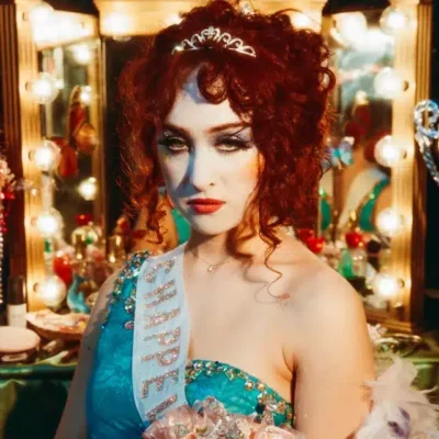 A woman with curly red hair, wearing a tiara and a blue, bejeweled dress with a sash, stands in front of a decorated vanity mirror with lights.
