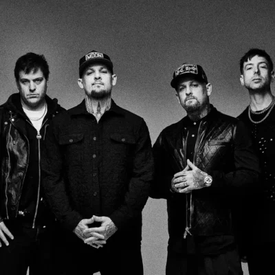 Four men stand side by side against a plain background, wearing dark clothing and hats, facing the camera with neutral expressions.