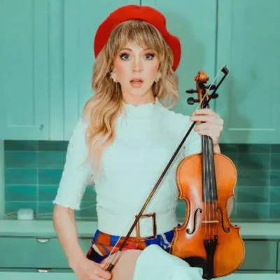 A woman wearing a red beret and white top sits in a kitchen holding a violin and bow, looking directly at the camera.