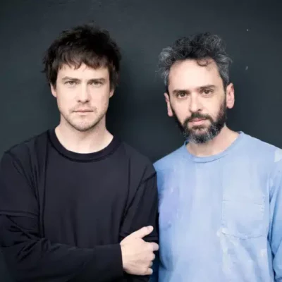 Two men stand side by side against a dark wall. One wears a black long-sleeve shirt, and the other wears a light blue shirt. Both look directly at the camera with neutral expressions.