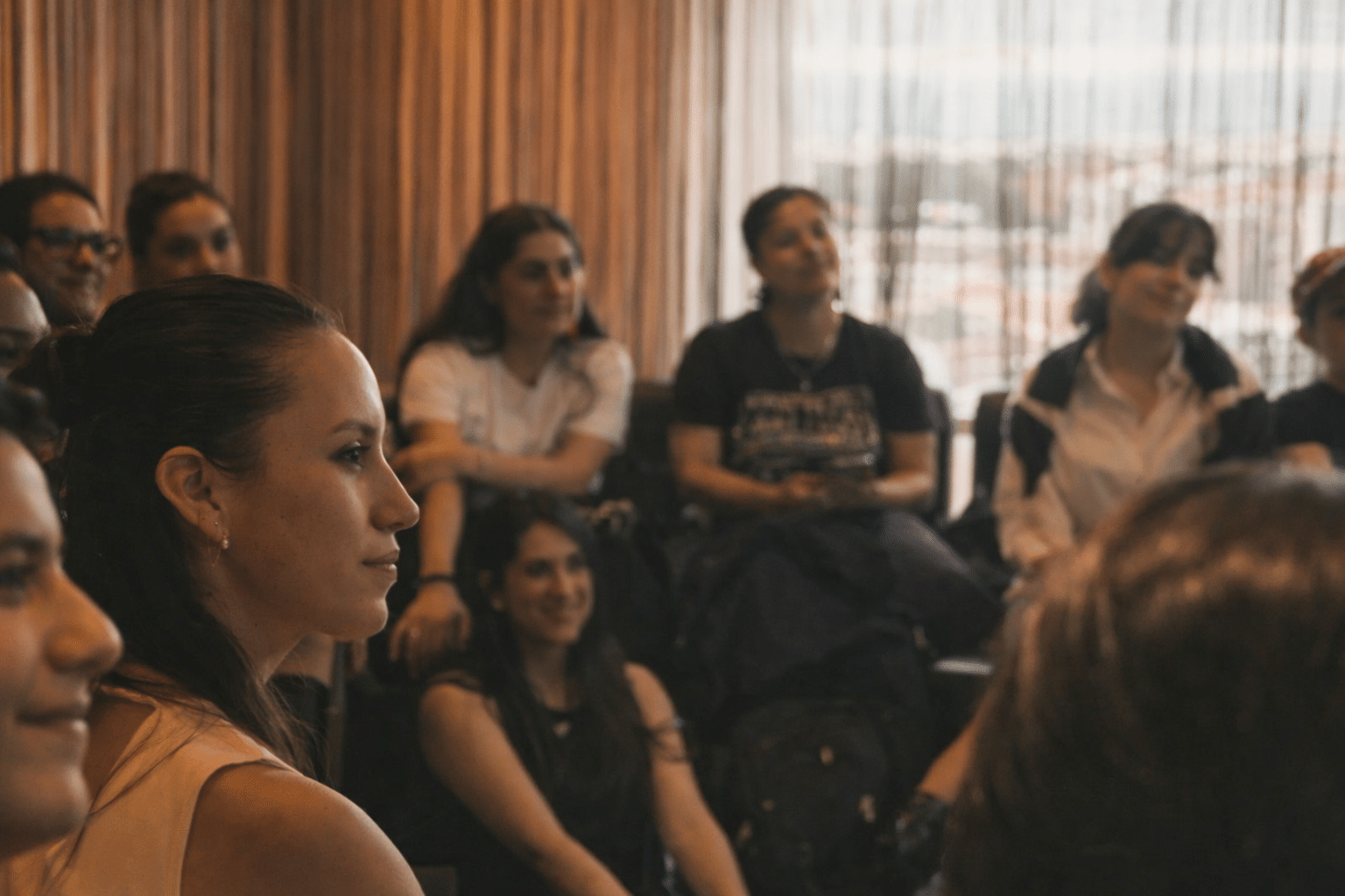 A group of Women Empowered sit indoors, attentively listening to someone outside the frame; soft daylight streams through sheer curtains in the background.