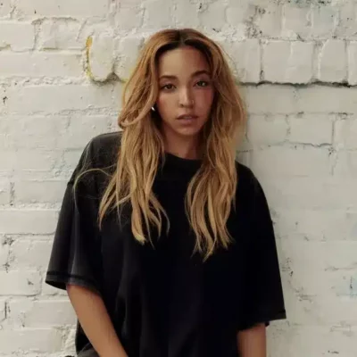 A woman with long, wavy blonde hair wearing a loose black t-shirt stands in front of a white brick wall, looking at the camera.