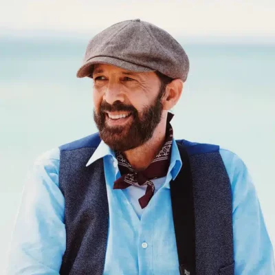 A bearded man wearing a flat cap, blue shirt, dark vest, and bandana around his neck stands outdoors with a blurred light blue background, embodying the creative spirit found in stories About Symphonic.