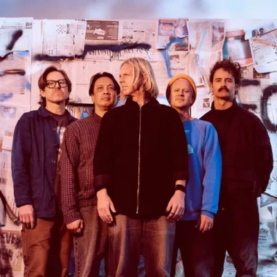 Five men stand in front of a wall covered with newspapers and graffiti, posing for a group photo—about Symphonic and their bold, urban vibe.