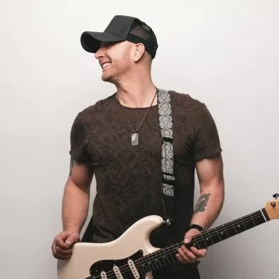 Man wearing a black cap and patterned t-shirt smiles while holding an electric guitar against a plain white background, capturing the creative spirit of About Symphonic.